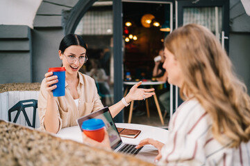 Astonished female colleagues communicate during friendly distance working at modern laptop computer, happy freelancers discussing business ideas for creating project planning collaborating at sidewalk