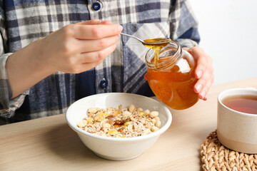 Woman putting honey into bowl with muesli at wooden table, closeup