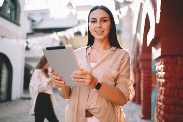 Half length portrait of pretty female with modern digital tablet in hands posing at urbanity, successful Caucasian blogger 20 years old with touch pad smiling at camera during sunny day in city