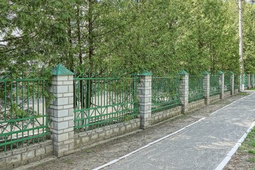 a long wall of green metal and white brick fence on the street in front of a row of coniferous trees