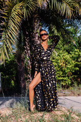 Fashion exotic portrait of slim tanned fit short dark hair woman with bronze make up in black golden long cape by palm tree, shadows falling on face and body from palm leaf. 