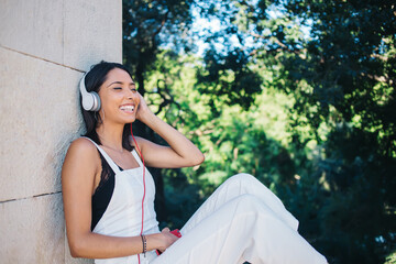 Cheerful positive woman millennial brunette excited with music in headphones and perfect sound, smiling caucasian 20s hipster girl listening playlist song entertaining on free time outdoors on weekend