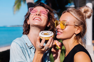 Two happy fit woman in pink and yellow sunglasses smiling having fun laugh with donuts outside villa hotel at natural day light.   