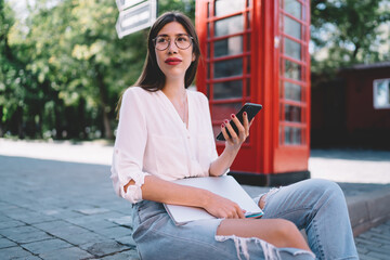 Beautiful hipster girl in optical eyewear resting at urban setting in London city sitting near red phone box and making online communication via cellular gadget, Hispanic woman using smartphone