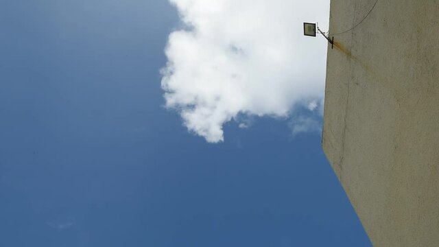 White clouds floating by on a deep blue sky framed with a building side wall and security flood light