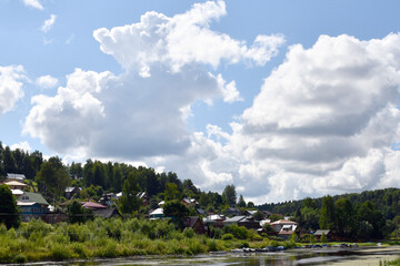 Fototapeta na wymiar Plyos, Ivanovo region, Russia, August 2020. A view of small houses among large green trees on the banks of a quiet river. Sun-lit large white clouds in the sky.