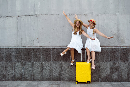 Beautiful Two Female Tourist Twins Are Sitting Near A Yellow Suitcase With Their Arms Spread Out To The Sides Imitate Flight. F