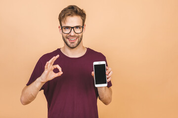 Close up portrait of a satisfied young man in t-shirt showing blank screen mobile phone isolated over beige background.