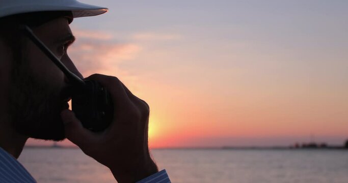 A seaport worker puts on a white helmet and talks on the radio. A seaport worker manages logistics by radio communication  on the background of the sea early in the morning.