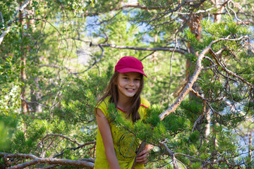 Naklejka na ściany i meble A smiling girl in a red cap sits on a pine tree in the forest in summer.