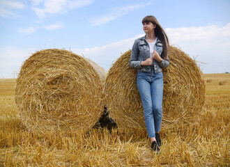 Naklejka na ściany i meble A young girl in denim clothes stands next to two bales of hay.