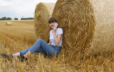 Naklejka na ściany i meble A young girl is on the phone, sitting on a harvested field of wheat, leaning back against large bales of straw.