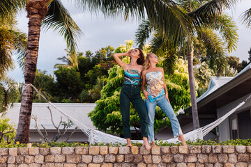 Two happy woman friends with sunglasses on vacation in tropical country. Females in top and jeans having fun at villa  hotel, palm trees on background.  
