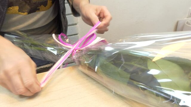 A woman florist in a flower shop ties a ribbon bouquet of roses in wrapping film for sale.