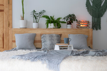 Fototapeta na wymiar Grey pillows on wooden double bed with green plants and mug of coffee. Bright interior of stylish room in rural cottage. Comfortable hotel. 