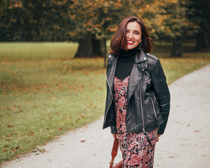 Smiling female dressed boho fashion style colorful long dress with black leather biker jacket with...