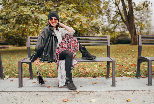 Sincerely laughing female dressed fashion boho style colorful long dress, warm knitted sweater with black leather biker jacket and Beanie Hat with flap bag sitting on bench at autumn city park