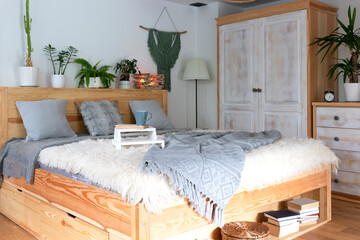 Stylish bedroom with wooden bed and wooden furniture. Cozy interior of room in hotel with grey pillows and plants. Bright and rural architecture.