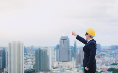 Asian male engineer or businessman Wearing a yellow helmet is standing on the rooftop of building and Point to the target in front of the tall building and cities