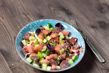 Salad with watermelon, cheese, basil leaves, sausage, cucumber, raddish, pepper, olive oil in blue bowl with fork on wooden table