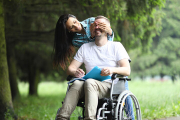Fototapeta na wymiar Happy woman stand behind and cover man eye with her palm. Cheerful man sit in wheelchair and hold notebook with pen in his hand.