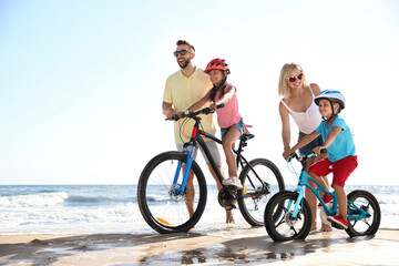 Happy parents teaching children to ride bicycles on sandy beach near sea