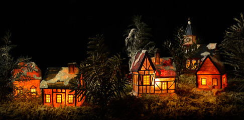 Panoramic scene of miniature Christmas countryside with lit tiny ceramic houses. Realistic homemade Xmas model of night village with trees, fake snow on roofs and Christian church on black background.