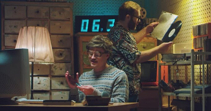 Caucasian man in glasses with mustache sitting at vintage monitor computer and playing with diskettes in hands. Male on background taking out disk record to play. Friends nerds in retro interior.