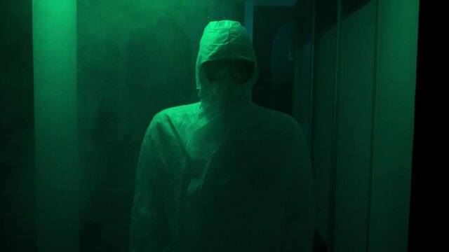 Man in protective suit and mask stands in dark. Stock footage. Creepy doctor in antiviral suit stands in dark corridor with green light. Dangerously high levels of infection