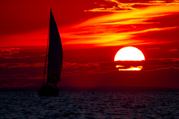 A spectacular sunset over Chesapeake Bay Maryland as captured from the east shore. Image features the large sun going down among clouds on a red sky and the silhouette of a sailboat with full sails.