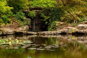 Fototapeta na wymiar A long exposure image from a decorated backyard pond with water lilies and a small waterfall where water flows over smooth rocks into the pool. The area is shaded covered with trees, moss.