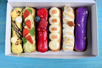 Set of delicious eclairs with glaze, raspberry and sugar decor in white paper box on blue wooden background. Delicious eclairs with berries and colorful topping 