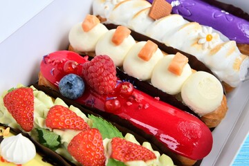 Set of delicious eclairs with glaze, raspberry and sugar decor in white paper box on blue wooden...