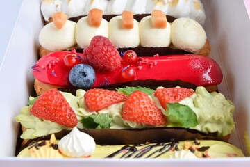Tasty French eclairs with icing, cream, fresh berries and sugar decor elements. Set of delicious eclairs with creative colorful decor in white paper box. 