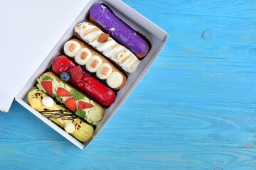 Set of delicious eclairs with glaze, raspberry and sugar decor in white paper box on blue wooden background. Delicious eclairs with berries and colorful topping 