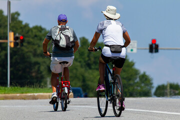 A senior couple wearing casual shorts and t shirts as well as hats is riding bikes near a highway on a sunny summer day. Man has a foldable pedal bike and the woman has a mountain bicycle.