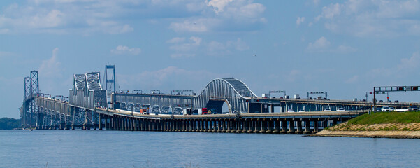 A day time wide angle image showing the rush hour traffic on Chesapeake Bay Bridge. It features detailed view of the bridge with columns and suspensions as well as the boats passing under.