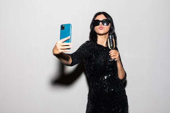 Young beautiful woman celebrating on party on white studio background, luxury dress, drinking champagne in glass, smiling, having fun, taking selfie photo on smart phone