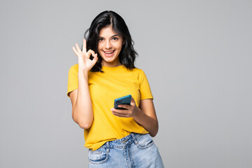 Attractive young girl showing ok gesture while holding blank screen mobile phone isolated over gray...