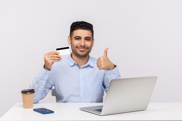 E-commerce. Elegant businessman sitting at workplace with laptop, holding credit card and showing thumbs up, recommending bank service, online purchase. indoor studio shot isolated on white background