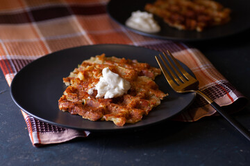 toasted Viennese zucchini waffles with sour cream sauce on two black plates, a checkered towel on a dark concrete background.