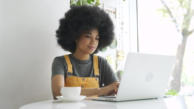 Happy stylish hipster gen z African American female student with afro hair using laptop computer, drinking coffee sitting at table at home, in cafe, remote studying, elearning or working in internet.