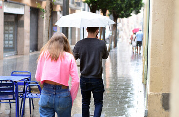 Young couple walking in the street under the rain. Boyfriend under an umbrella while girl is getting wet. 