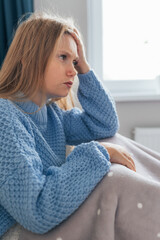 Beautiful blonde young woman having a strong headache sitting in bed. Female holding head with her hands, suffering on her face