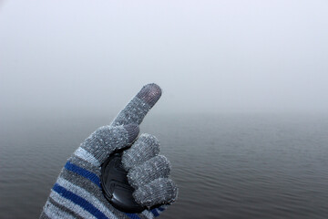 A man's hand in a woolen glove. In the palm of his hand the lid from the camera lens is clamped. There is a mist above water in the background.