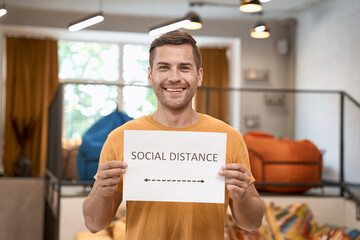 Reopening office safely. Young happy man showing paper with text SOCIAL DISTANCE at camera and smiling, standing in the modern coworking space