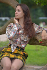slender brunette girl in a plaid skirt and blouse in the summer by a tree in the park