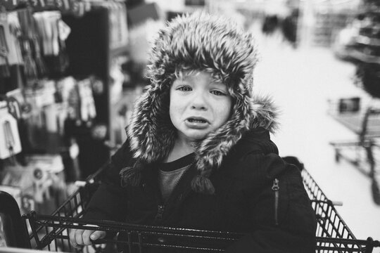 A little boy in a furry hat crying in a shopping cart.