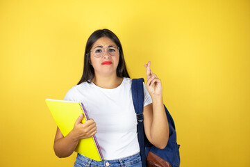 Young beautiful student woman wearing backpack holding notebook over isolated yellow background gesturing finger crossed smiling with hope