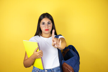 Young beautiful student woman wearing backpack holding notebook over isolated yellow background showing and pointing up with fingers number one while is serious